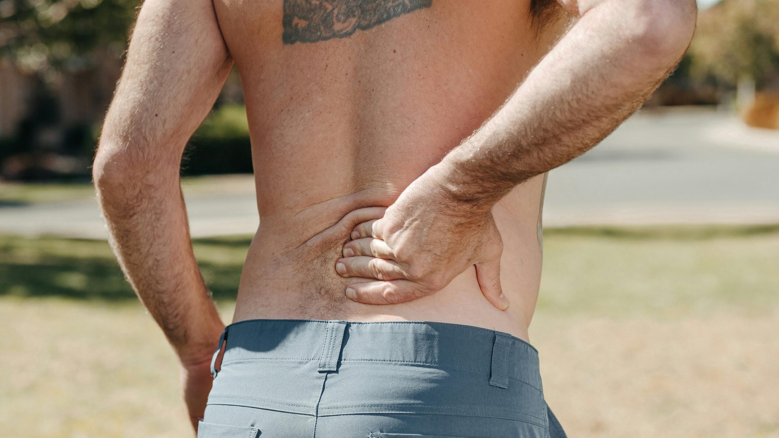 Shirtless man holding his lower back outdoors indicating pain or discomfort.